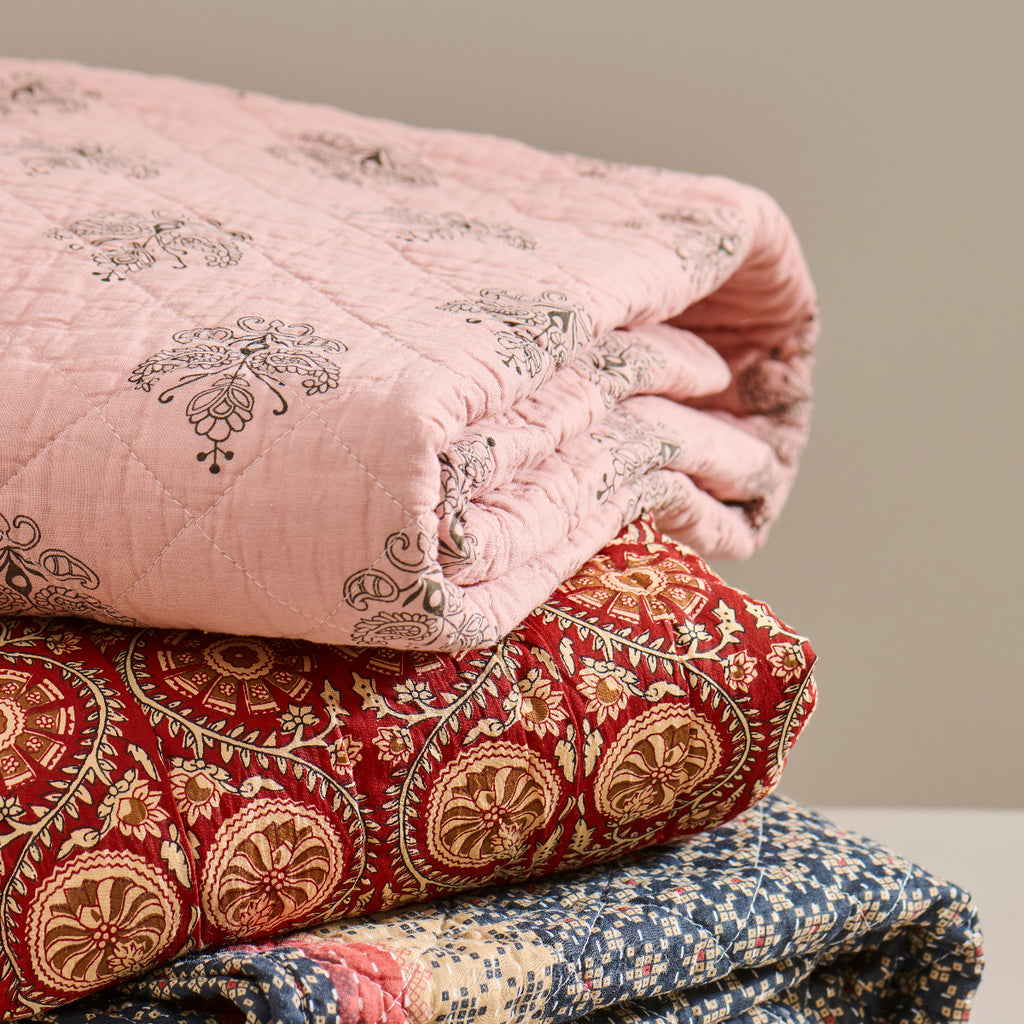 Stack of patterned quilts in pink, red, blue, and gray on a neutral background