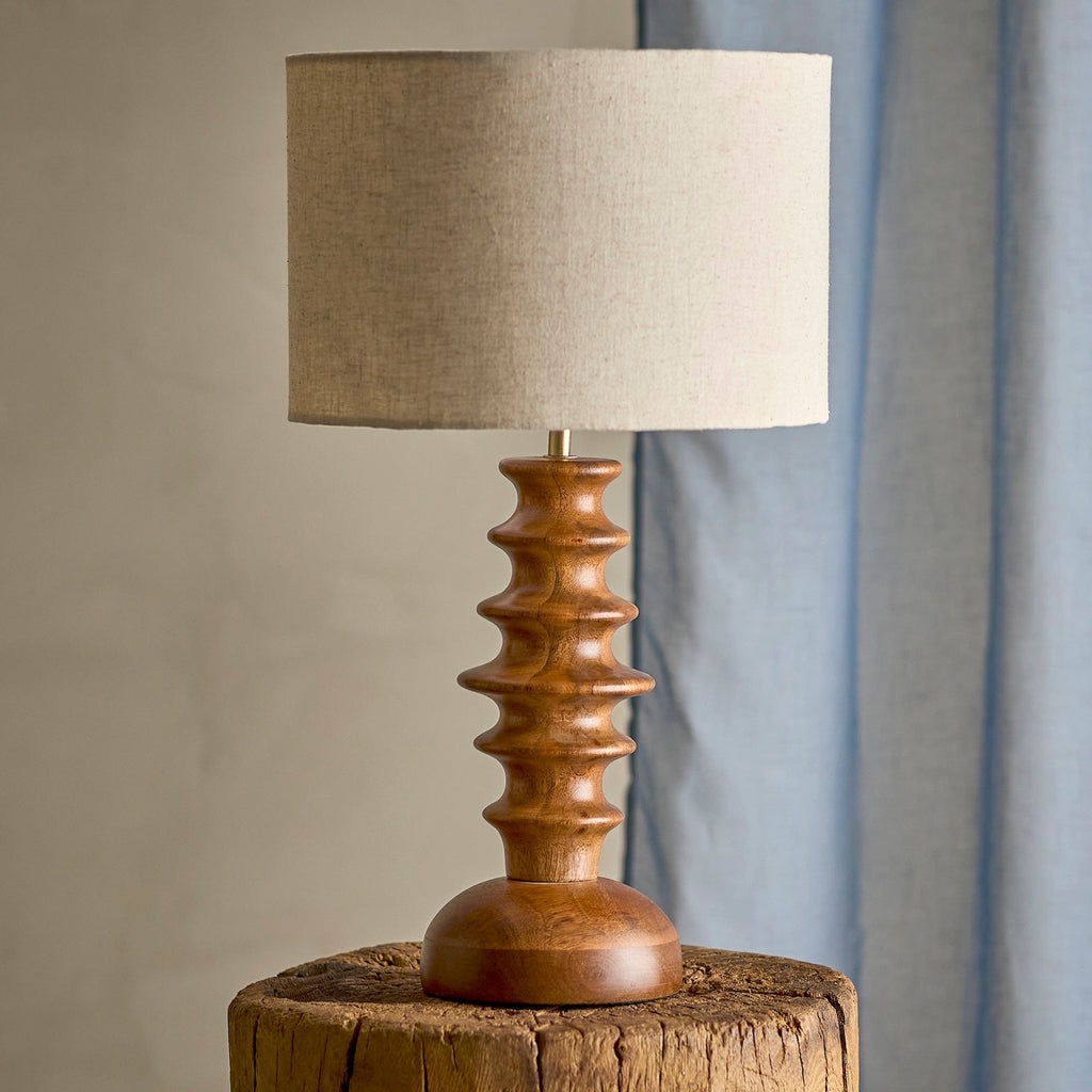 A table lamp with a sculptural, ribbed wooden base and a beige linen drum shade, displayed on a rustic wooden stool against a neutral background with a blue curtain.