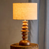 A table lamp with a warm glowing linen drum shade and a sculptural, tiered wooden base, displayed on a rustic wooden stool against a neutral gray background.