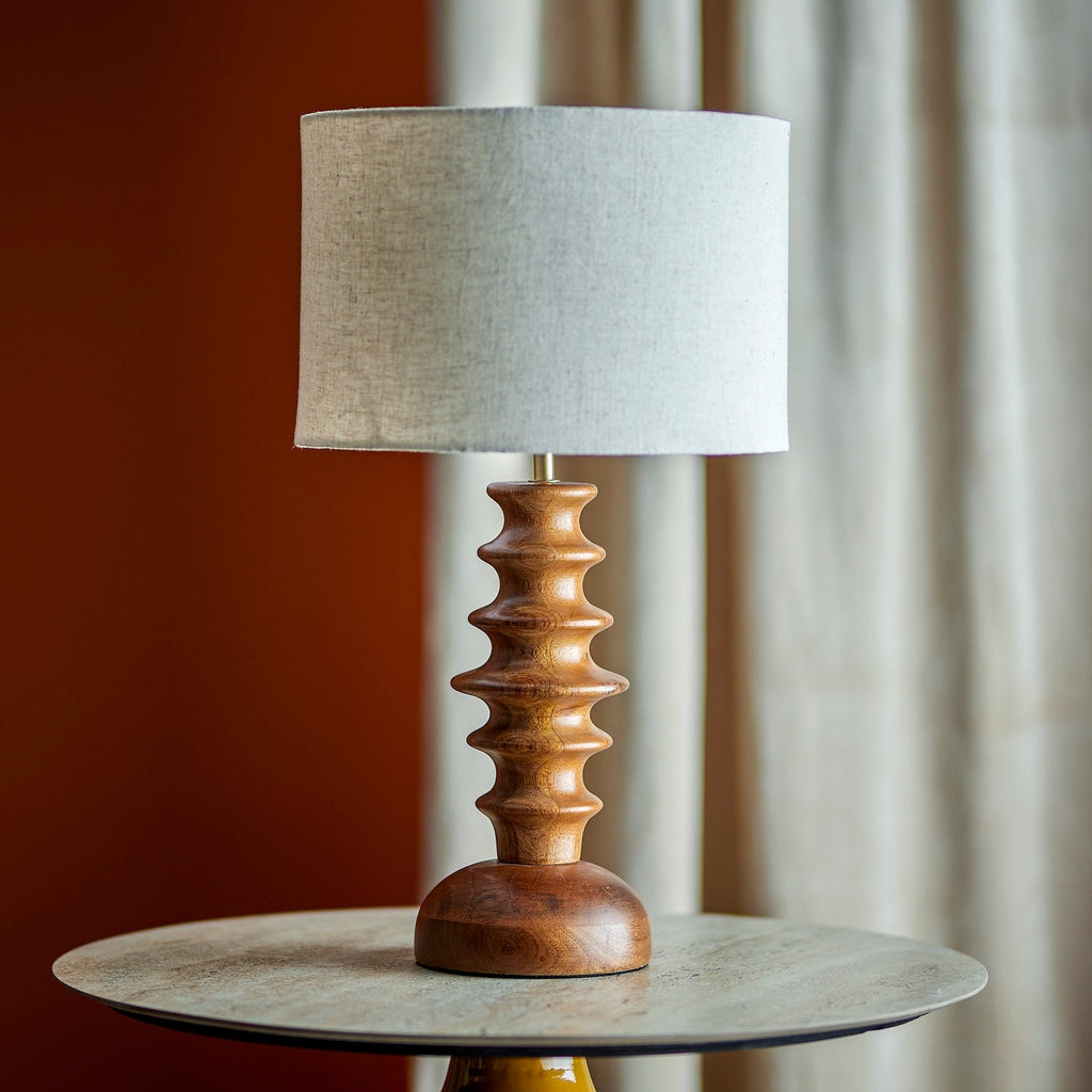 A table lamp with a textured light gray fabric drum shade and a sculptural, ribbed wooden base in a warm medium brown, displayed on a round side table.