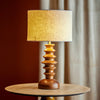 A mid-century modern table lamp with a sculptural stacked wooden base and a textured beige linen drum shade, illuminated and set on a round wooden side table against a warm brown and beige curtain background.