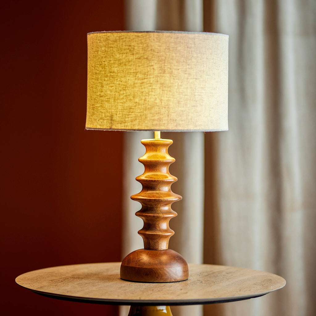 A mid-century modern table lamp with a sculptural stacked wooden base and a textured beige linen drum shade, illuminated and set on a round wooden side table against a warm brown and beige curtain background.