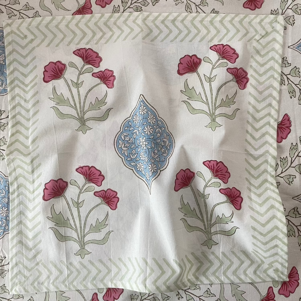 A square white fabric napkin or placemat featuring a central blue and gold ornamental motif, surrounded by four pink floral clusters with green stems, and a green-and-white chevron border. It is laid flat on a patterned tablecloth.